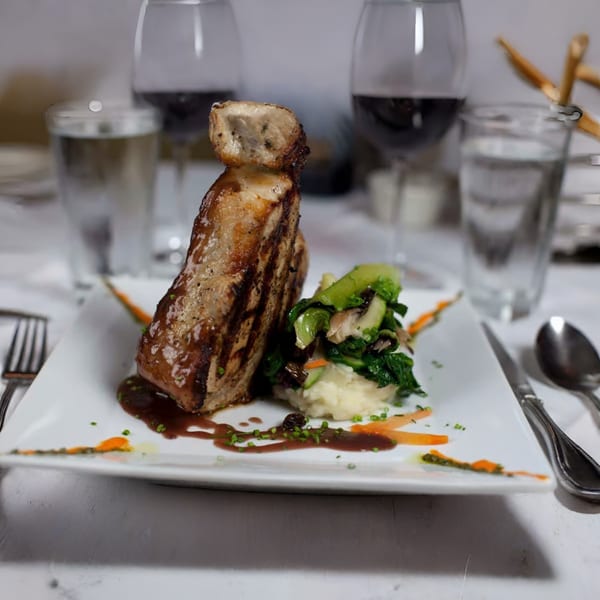 Grilled Italian pork chop with mashed potatoes, greens, and red wine on a white plate.