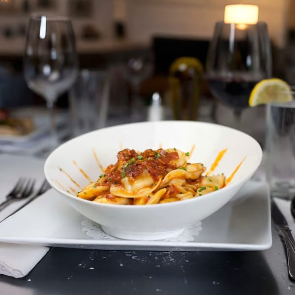 A white bowl of ravioli with red sauce and chives, set on a table with wine glasses.